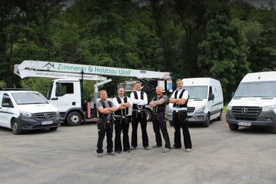 Fünf Zimmerer der Zimmerei Wolf in Zunftkleidung stehen nebeneinander vor dem Kranwagen und den Firmenfahrzeugen.