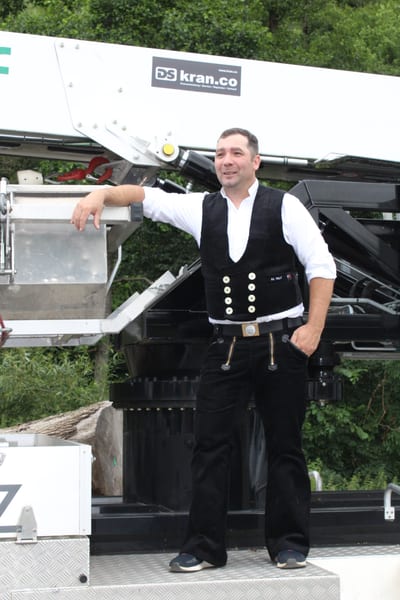Manfred Wolf in Zimmerer-Zunftkleidung mit Weste und Schlaghose, angelehnt an den Kranwagen des Betriebs.